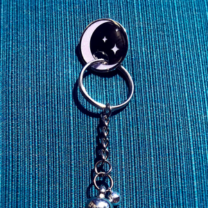 Sparkly lavender and silver moon charm pin with keyring hanging from it on turquoise cloth background.