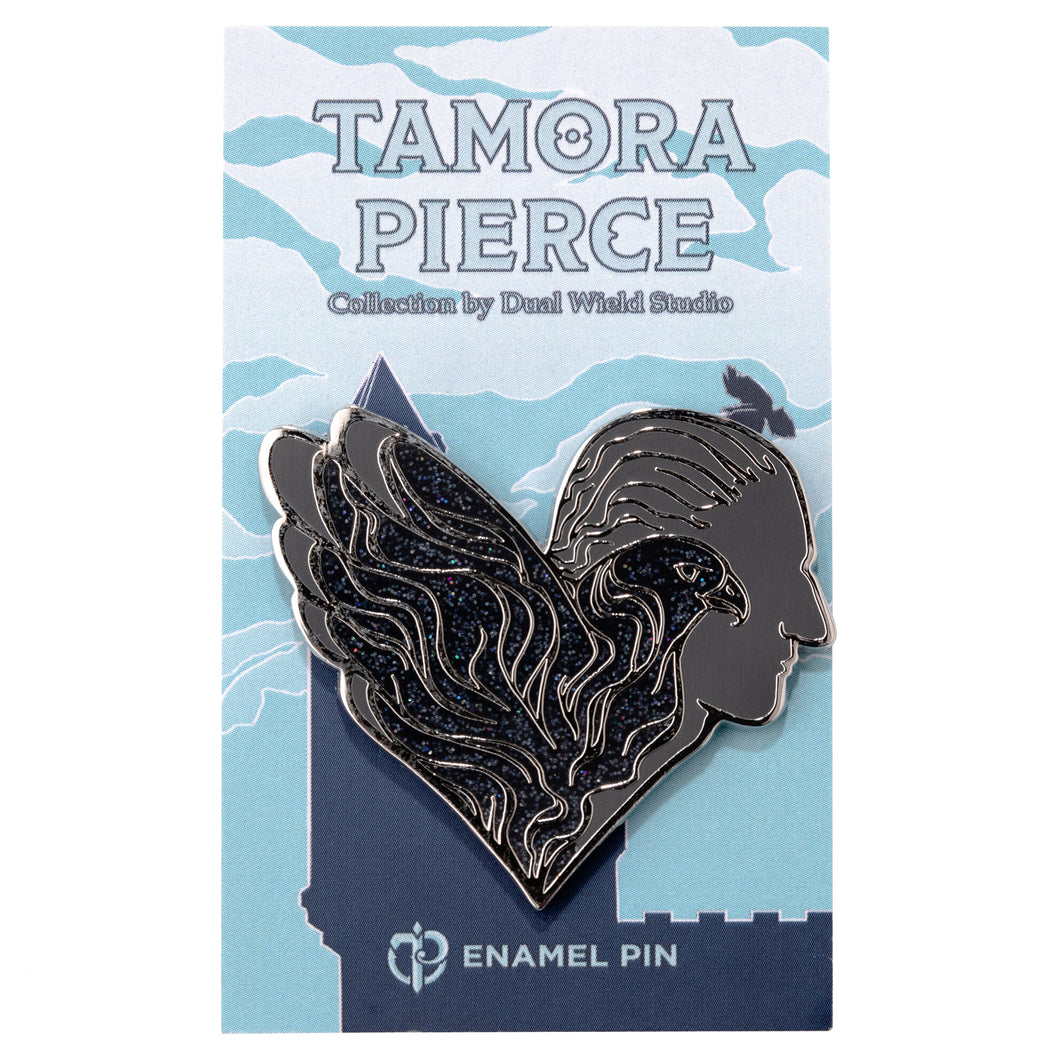 Glittering black and grey enamel pin that shows a hawk in profile, and Numair in profile behind it. The whole pin evokes a heart shape and is pinned to a light blue castle Tamora Pierce backing card.