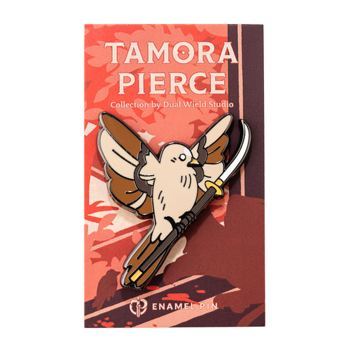 An enamel pin of a tan and brown sparrow in flight, holding a glaive weapon. Pin is attached to a coral and brown Tamora Pierce backing card.