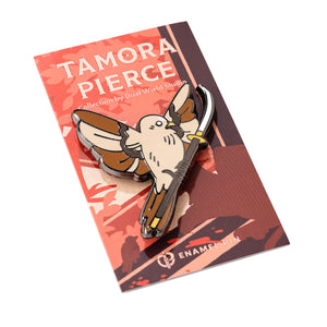 Angled view of Keladry Crown Sparrow Pin. The enamel pin is a tan and brown sparrow in flight, holding a glaive weapon. Pin is attached to a coral and brown Tamora Pierce backing card.