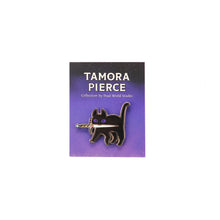 Load image into Gallery viewer, Purple-eyed black cat with gold enamel pin. Cat is standing and holding white, black, and gold dagger in its mouth and is pinned to starry purple and black Tamora Pierce backing card.