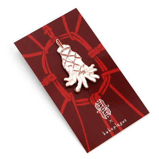 A rose gold pin depicting hands tenderly tied with rope on red backing card with white background.