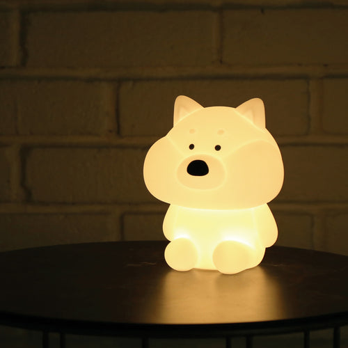Shiba mood lamp softly brightens are dark room on a bedside table in front of white bricks.