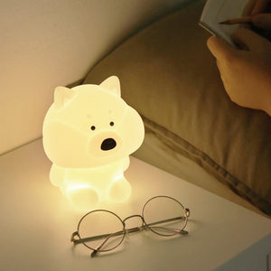 Model's hands are shown writing in bed beside shiba mood lamp and glasses on a white bedside table.