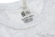 Load image into Gallery viewer, Closeup of printed-on-fabric tag with Dual Wield Studio x Tamora Pierce logos above care and wash instructions.