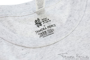 Closeup of printed-on-fabric tag with Dual Wield Studio x Tamora Pierce logos above care and wash instructions.